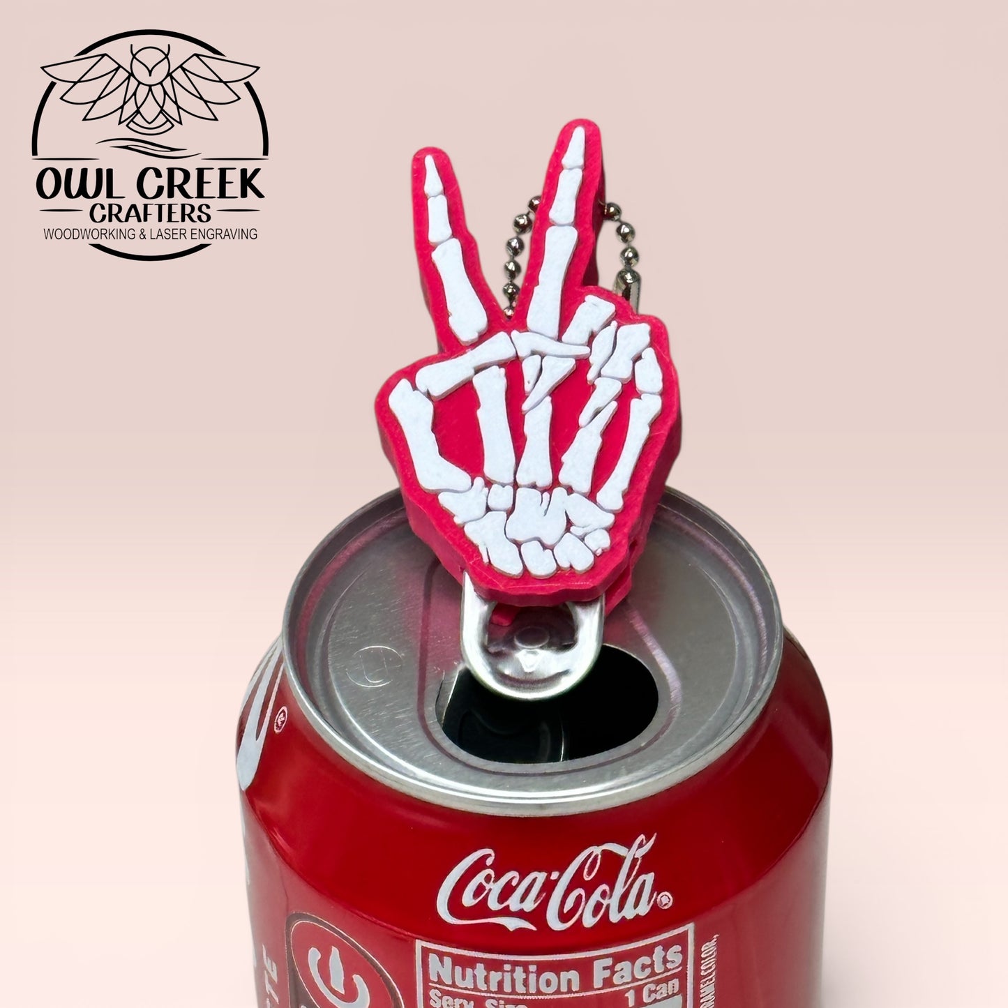 Magenta and white skeleton hand making a peace sign bottle opener on a Coca-Cola can with a beige background and an Owl Creek Crafters logo