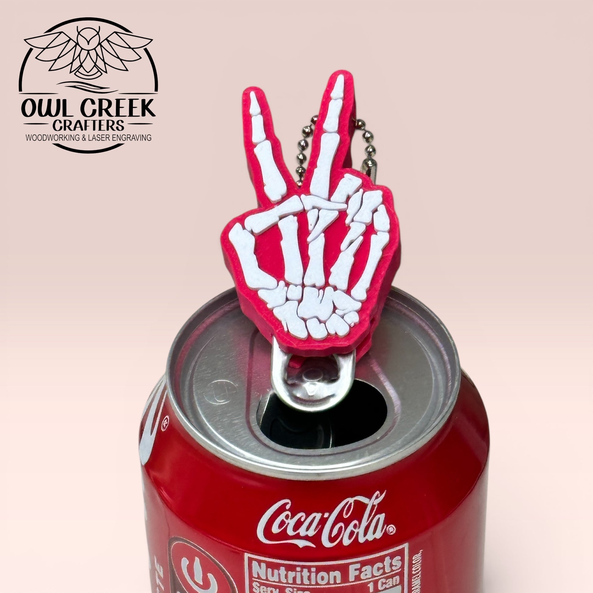 Magenta and white skeleton hand making a peace sign bottle opener on a Coca-Cola can with a beige background and an Owl Creek Crafters logo