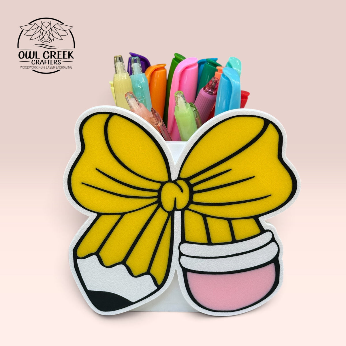 A 3d printed white pencil holder with a design in the front of a pencil in the shape of a bow. The pencil holder has colorful pens and highlighters in it. On a pink background with the Owl Creek Crafters logo in the top left corner