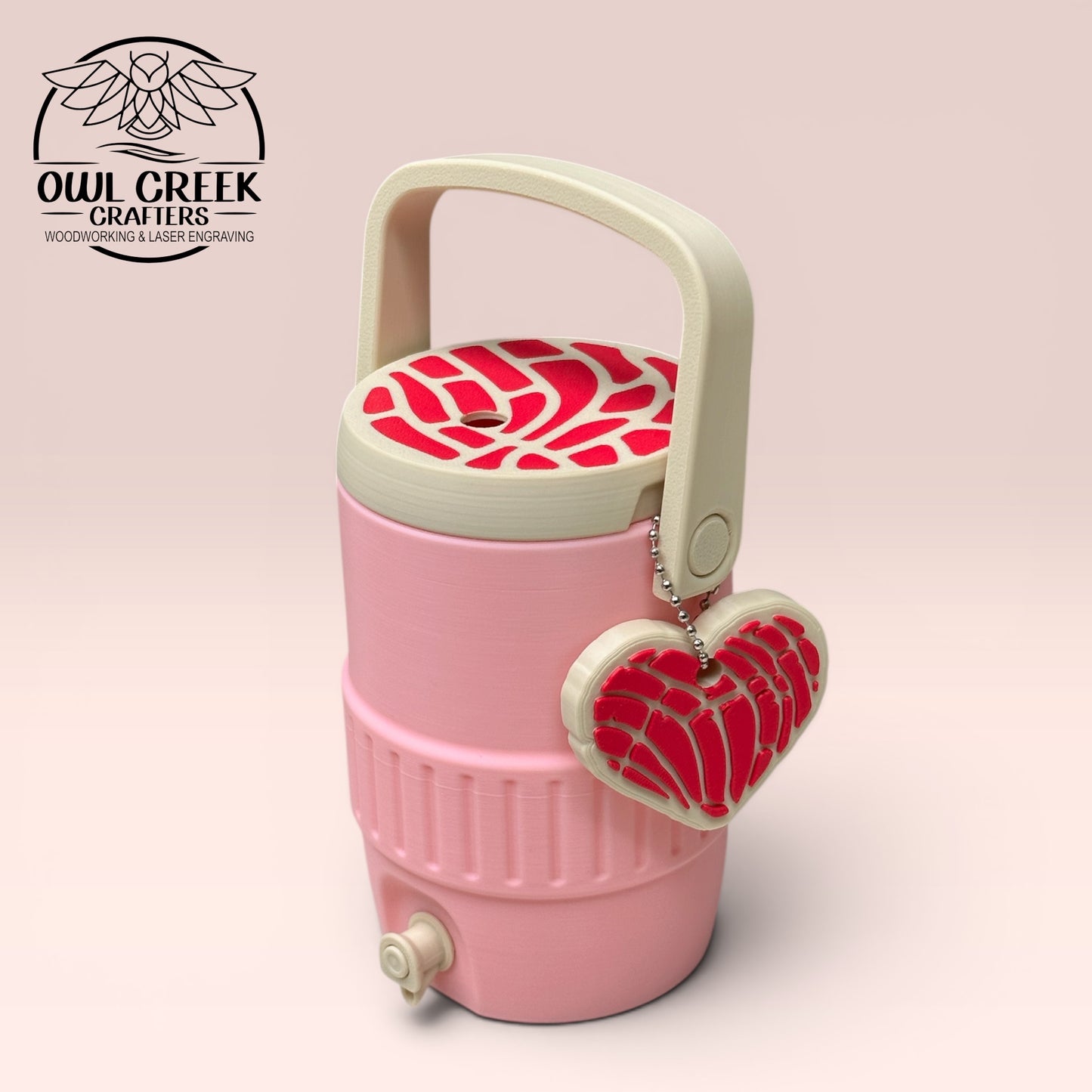 Pink can cooler coozie with a tan and  magenta lid and handle, and a tan and magenta heart shaped keychain, on a beige background with the Owl Creek Crafters Logo