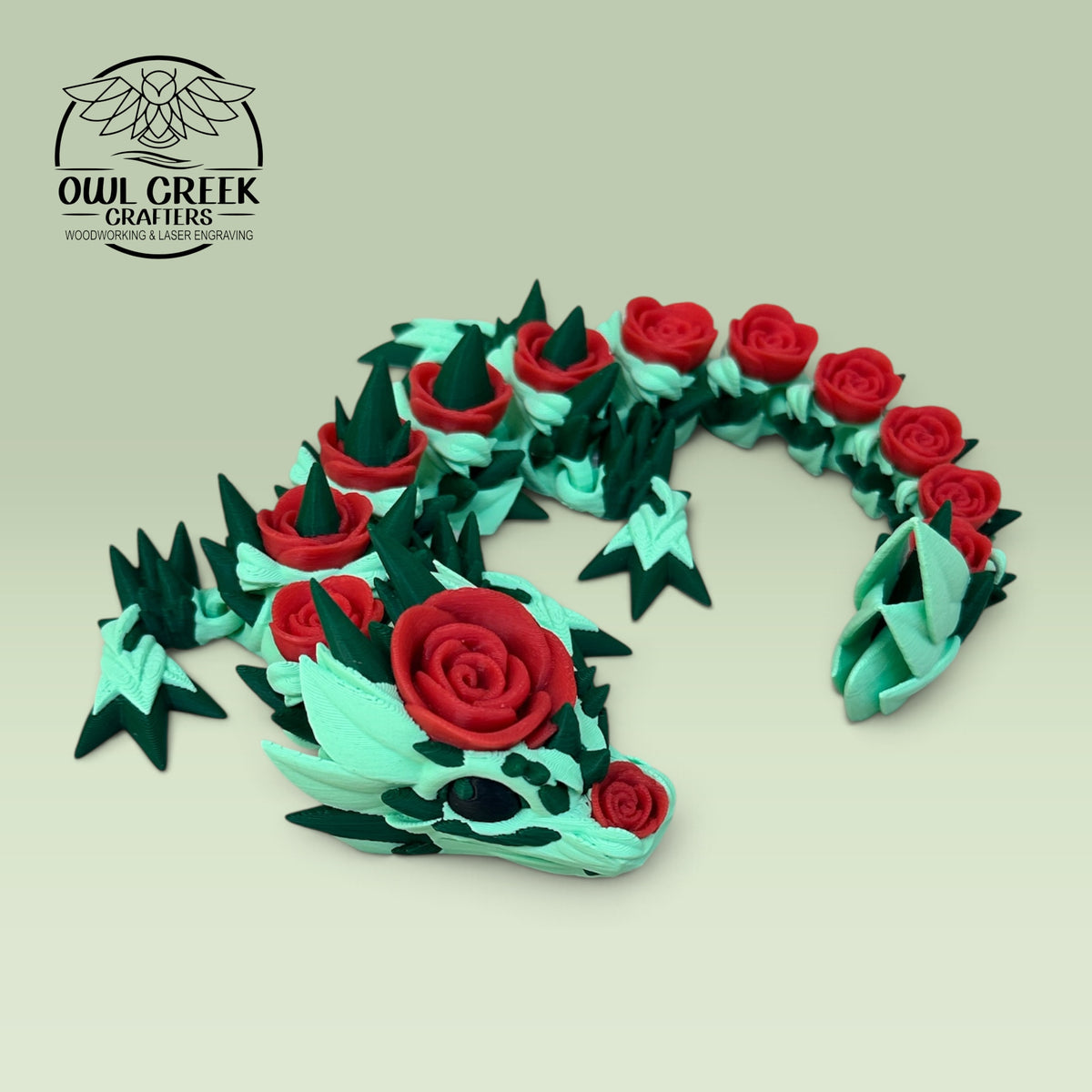 A 3D-printed articulated dragon figurine in mint green and dark green, decorated with red rose details, displayed on a light green background. The Owl Creek Crafters logo is visible in the top-left corner.