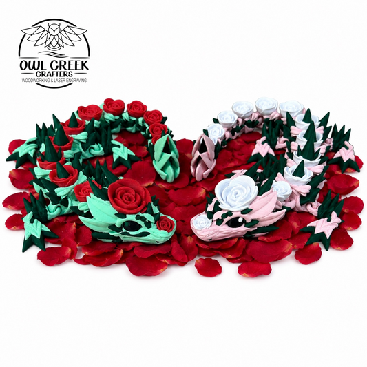 Two 3d printed rose dragons, one mint green with red roses and one light pink with white roses, displayed on a bed of red paper rose petals against a white background. The Owl Creek Crafters logo is in the top left corner.