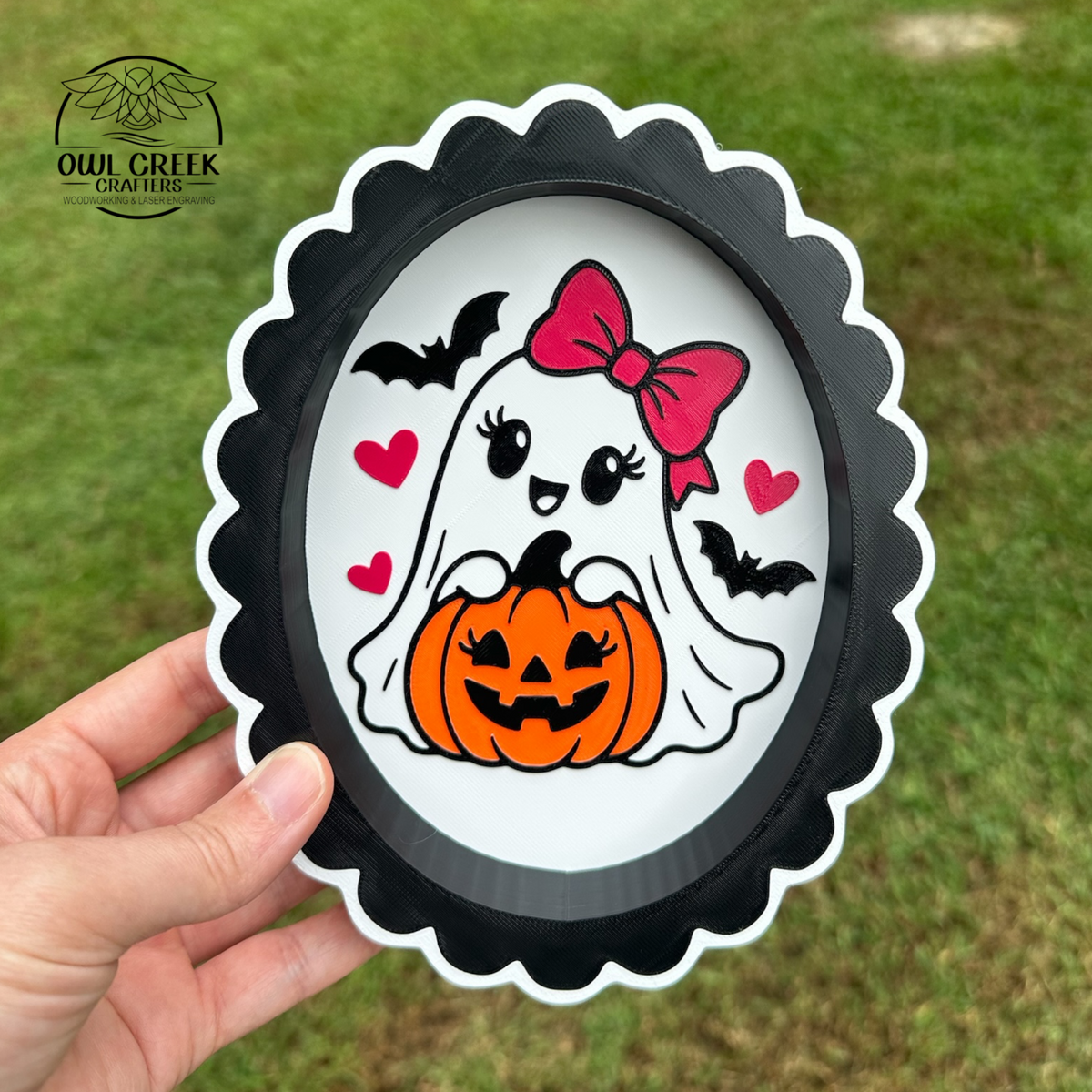 A hand holds a 3d printed scalloped oval trinket tray featuring a cute white ghost with a pink bow, holding an orange jack-o'-lantern, with black bats and hearts on a white background. The tray is photographed outdoors against a blurred green grass background. The Owl Creek Crafters logo is in the top left corner.