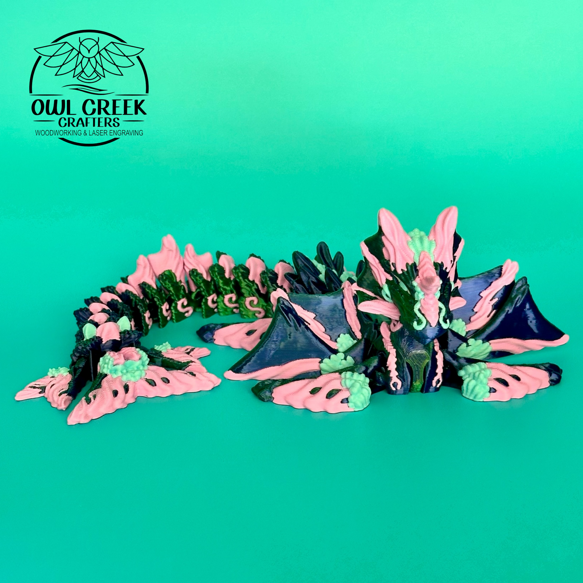 A 3d printed dragon figurine by Owl Creek Crafters, featuring pink, green, and blue colors, displayed on a solid green background, with a 'Owl Creek Crafters' logo in the top left corner