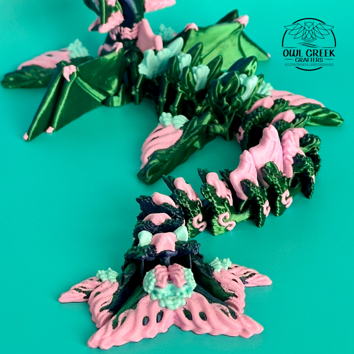 A close-up of a 3d printed fantasy dragon figurine in metallic green and pink, with a turquoise background and the Owl Creek Creations logo in the upper right.