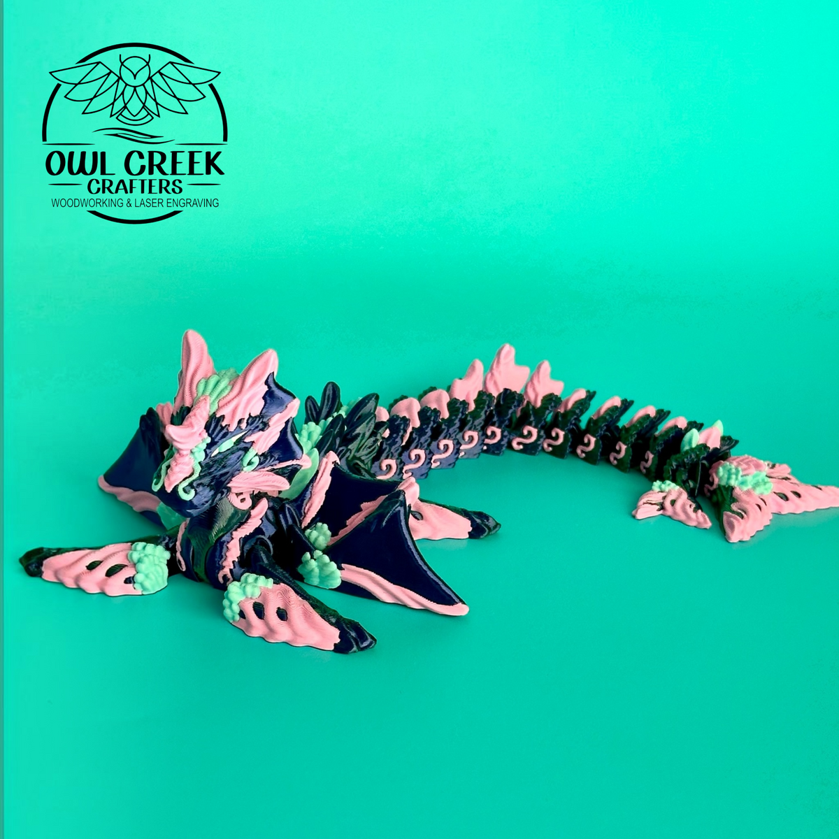 A 3d printed dragon figurine by Owl Creek Crafters, featuring pink, green, and blue colors, displayed on a solid green background, with a 'Owl Creek Crafters' logo in the top left corner