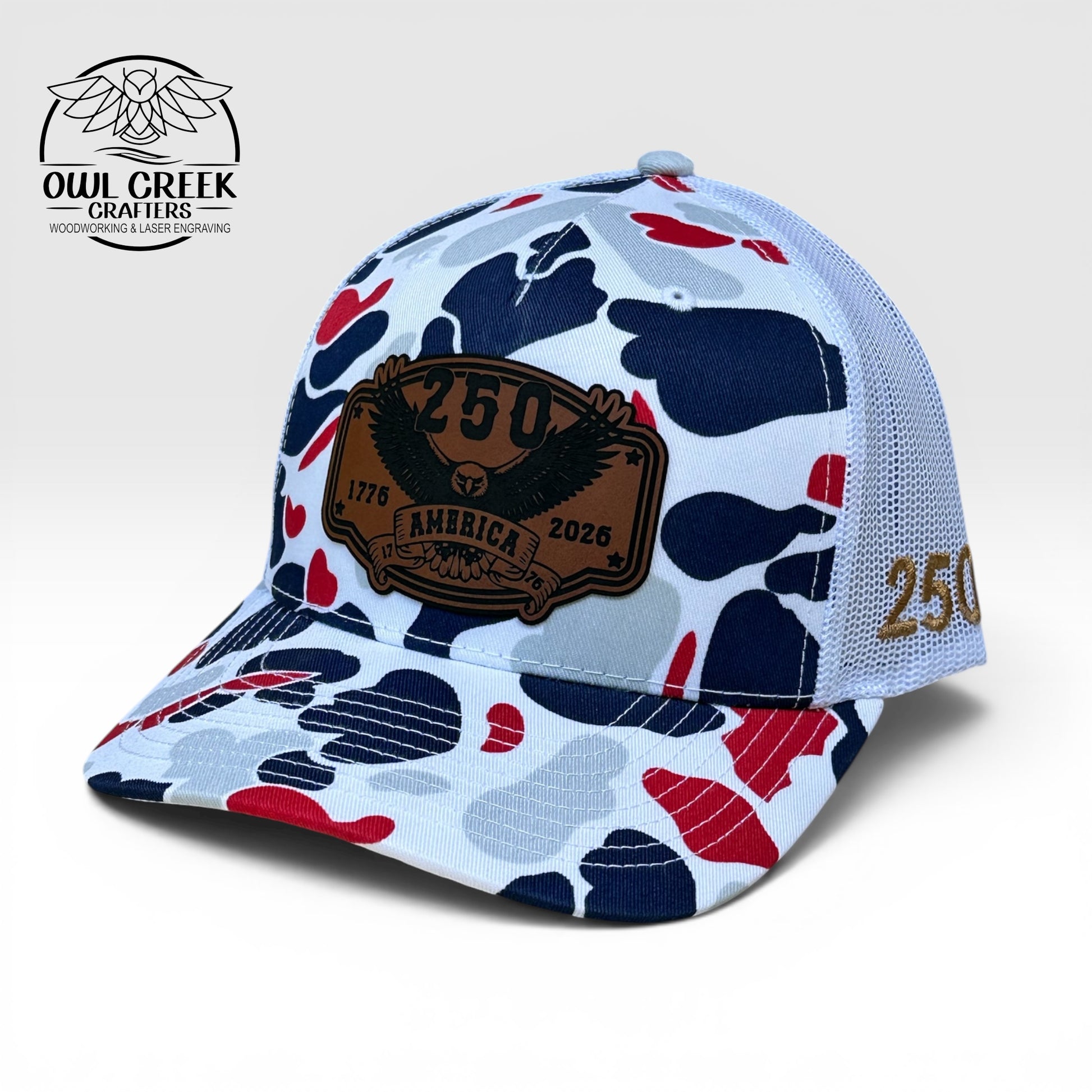 Red, white, and blue camouflage pattern hat with a leatherette patch that says "250 America, 1776-2026", and and embroidered 250 on the left side of the hat, with a light beige background and the Owl Creek Crafters Logo