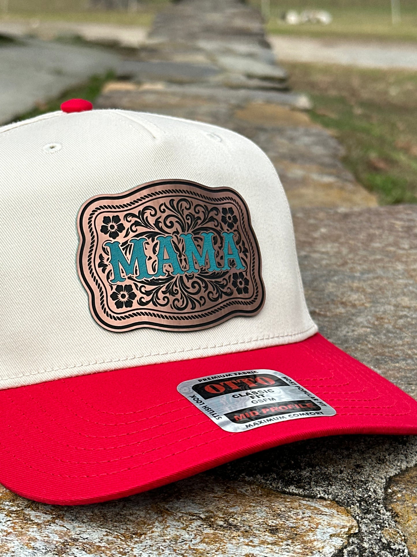 Close up of a two-tone cap with a white top and a red bill with a bronze acrylic patch with the text 'MAMA' in turquoise sitting on a stone wall.