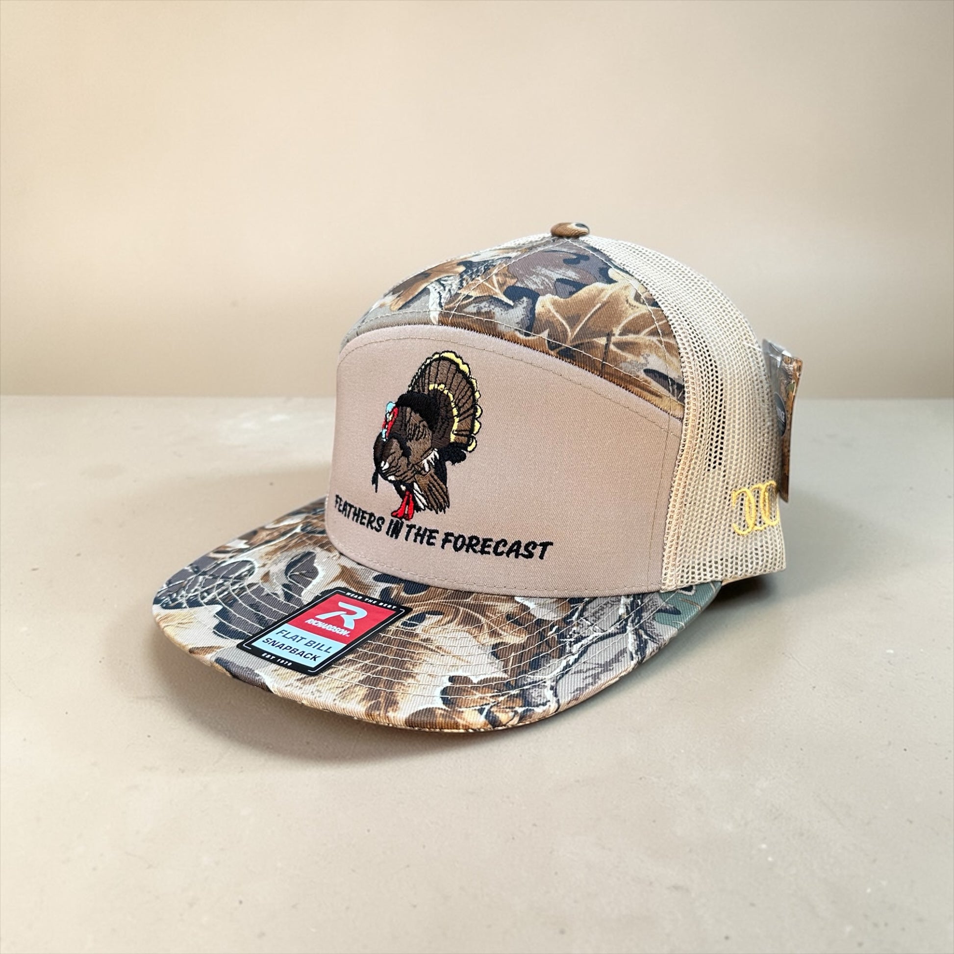 Camouflage and beige trucker cap with an embroidered turkey design and text Feather in the Forecast with a mesh back on a beige background