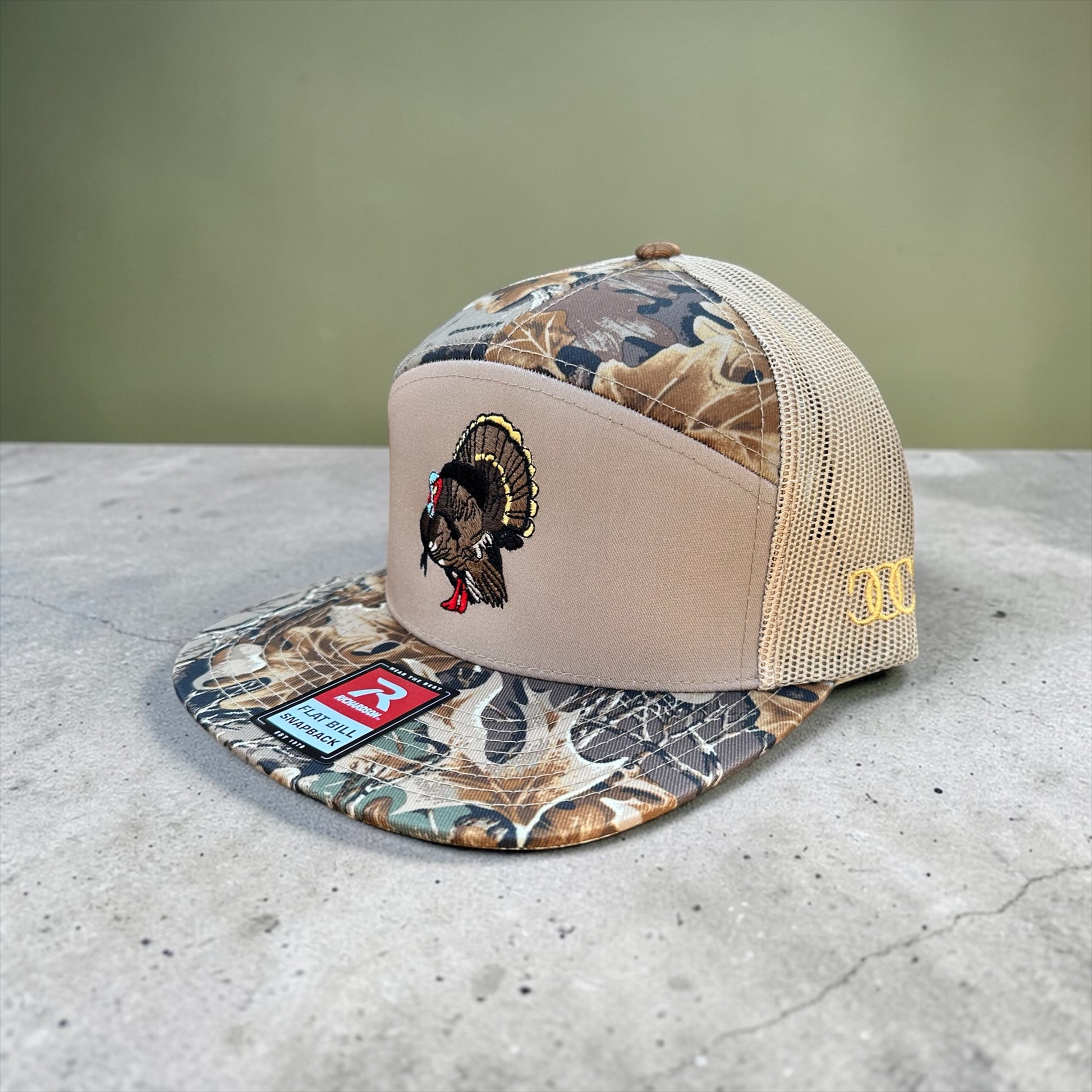 Camouflage and beige trucker hat with and embroidered turkey with mesh back and a visible brand logo on a concrete surface