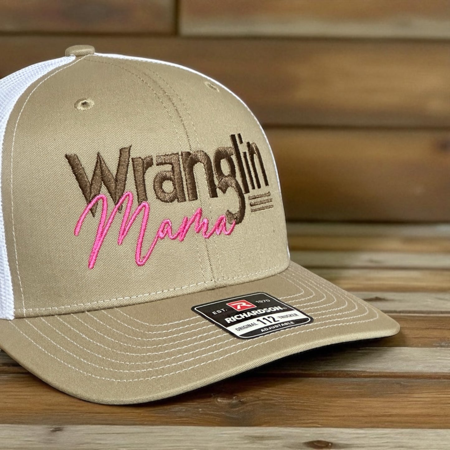 Beige trucker cap with 'Wranglin Mama' text and a white mesh back on a wooden surface