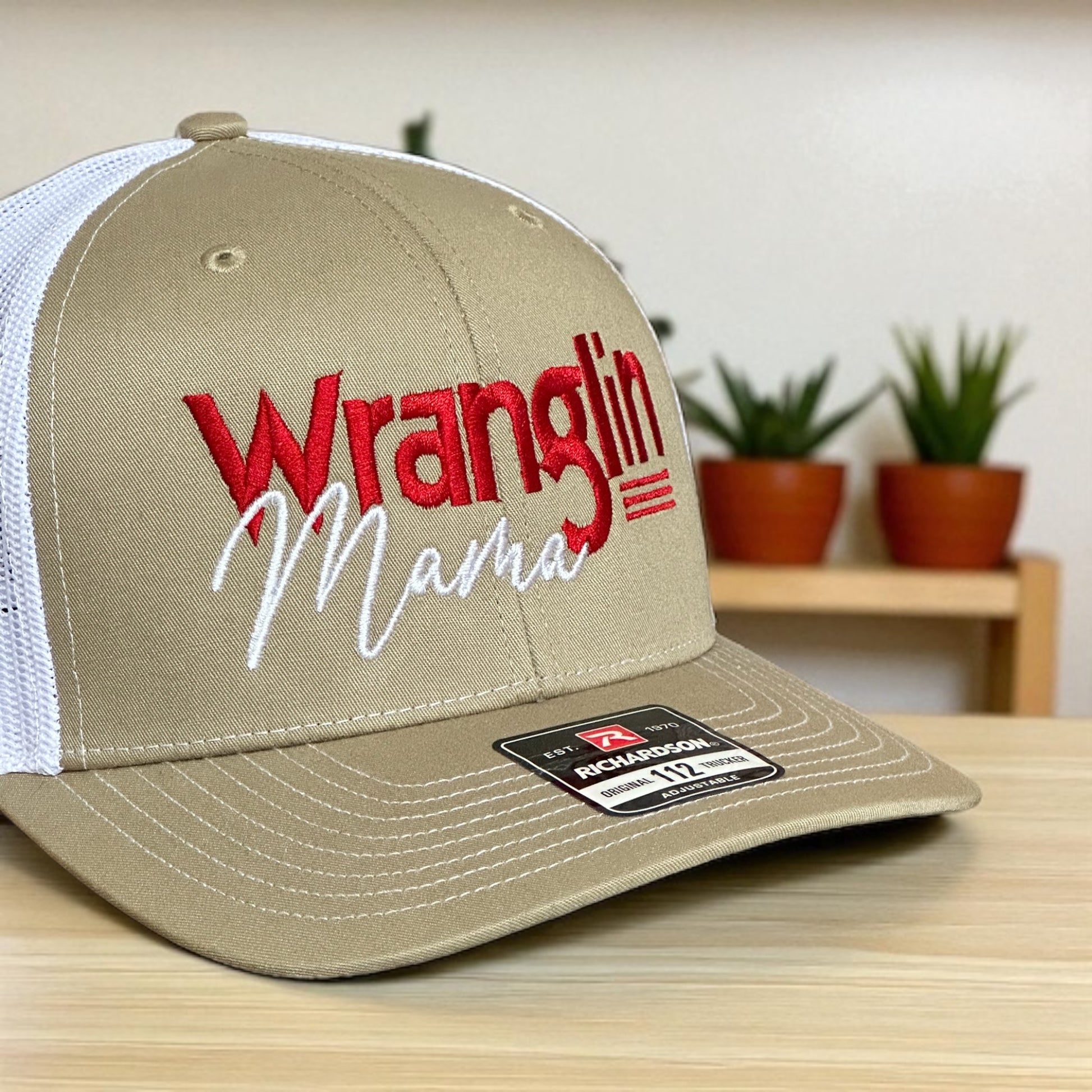 Beige trucker cap with embroidered 'Wranglin Mama' text, and a white mesh back on a wooden surface with plants in the background
