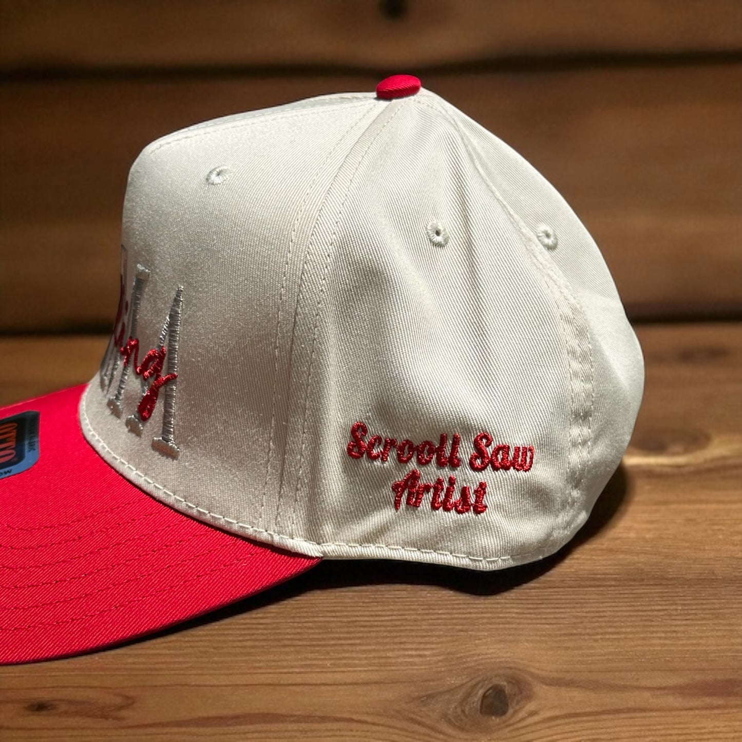 Two-tone cap with a light beige top and a red bill with the embroidered 'Scroll Saw Artist' text on the side of the cap, on a wooden surface