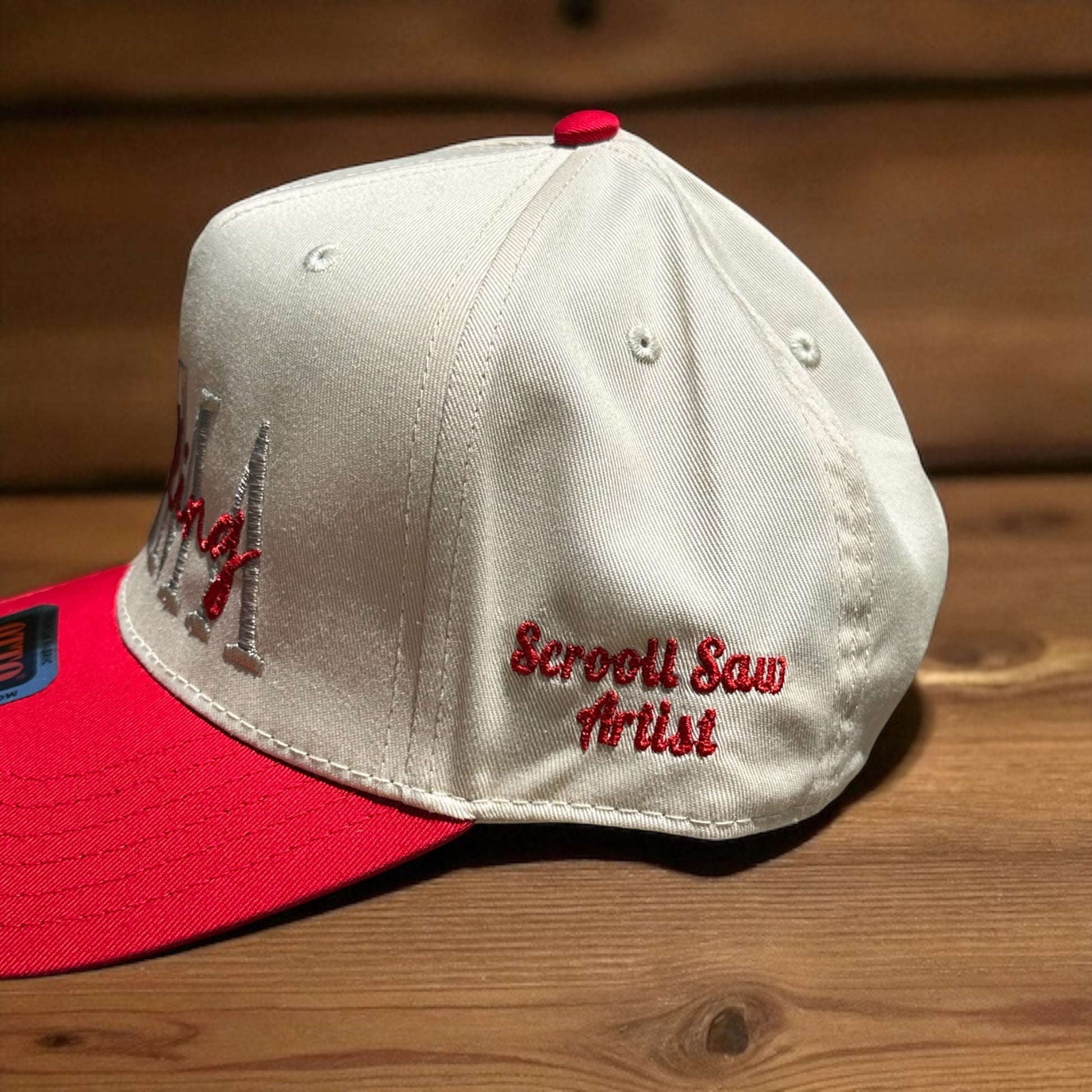 Two-tone cap with a light beige top and a red bill with the embroidered 'Scroll Saw Artist' text on the side of the cap, on a wooden surface