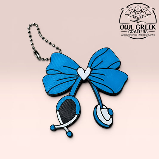 Blue stethescope bow keychain with a white heart in the center on a beige background, branded 'Owl Creek Crafters'.