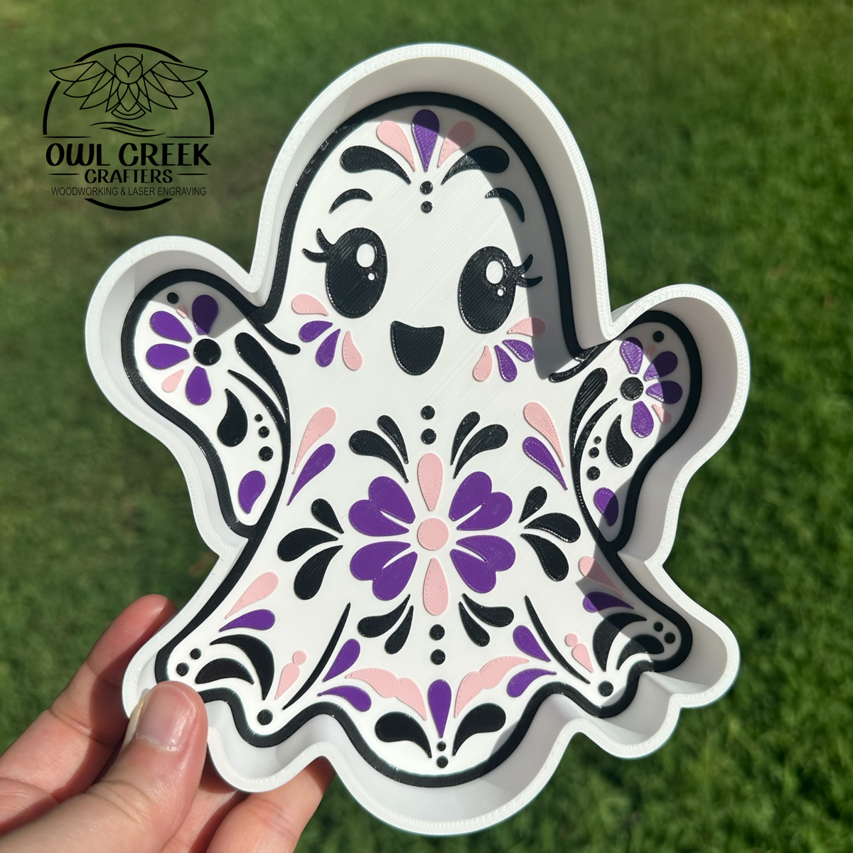 A hand holds a white ghost-shaped trinket tray decorated with a colorful Talavera pattern in purple, pink, and black, set against a green grass background. The Owl Creek Crafters logo is visible in the top left corner.