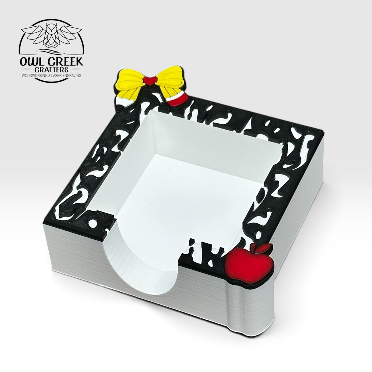 A square 3d printed sticky note holder, with a pencil bow, apple, and composition notebook design on  the top. On a light beige background with the Owl Creek Crafters logo in the top left corner.