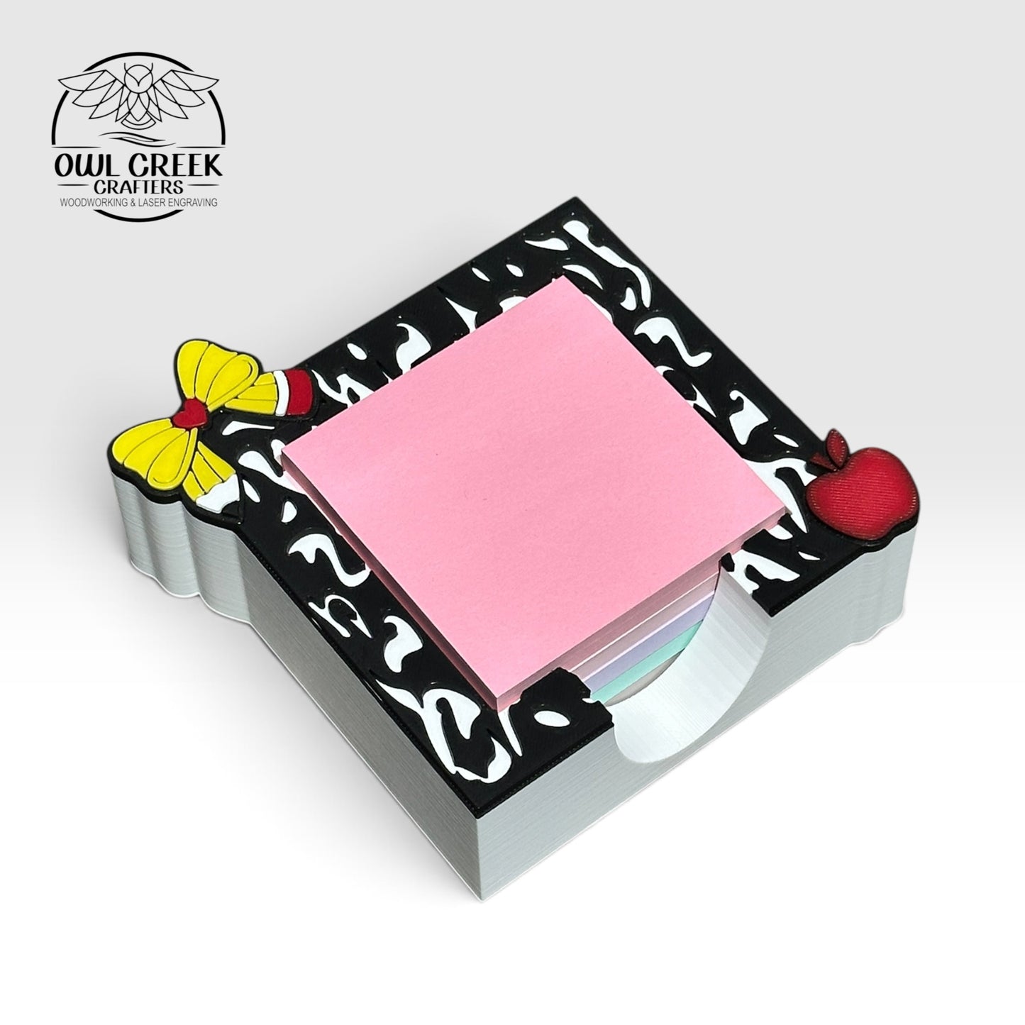 A square 3d printed sticky note holder, with a pencil bow, apple, and composition notebook design on  the top. Pink sticky notes in it. On a light beige background with the Owl Creek Crafters logo in the top left corner.