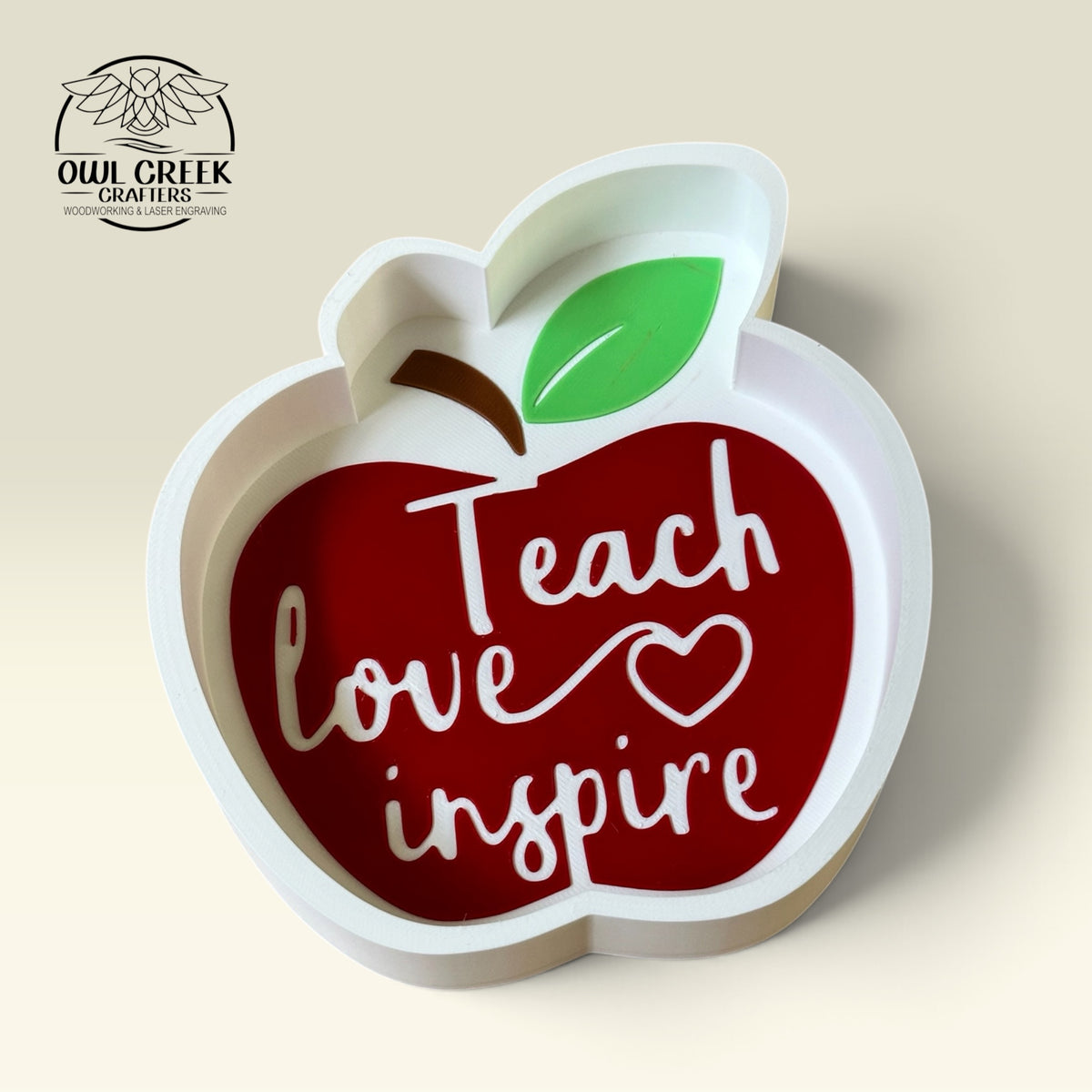 An apple-shaped decorative trinket tray with a white frame and a red apple in the center featuring the words 'Teach love inspire' in white script. The apple has a green leaf and a brown stem, and the sign is photographed on a light beige background, with the 'Owl Creek Crafters' logo in the top left corner