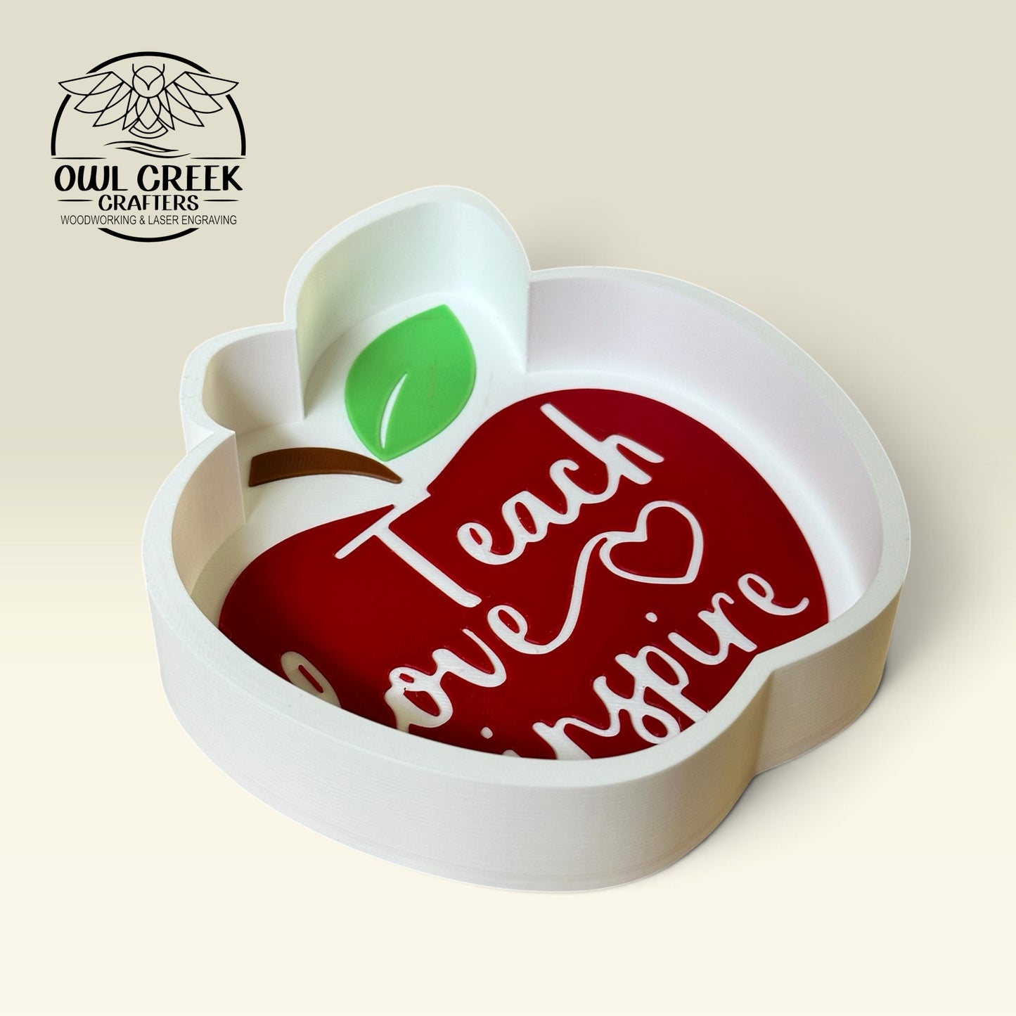 An apple-shaped decorative trinket tray with a white frame and a red apple in the center featuring the words 'Teach love inspire' in white script. The apple has a green leaf and a brown stem, and the sign is photographed on a light beige background, with the 'Owl Creek Crafters' logo in the top left corner