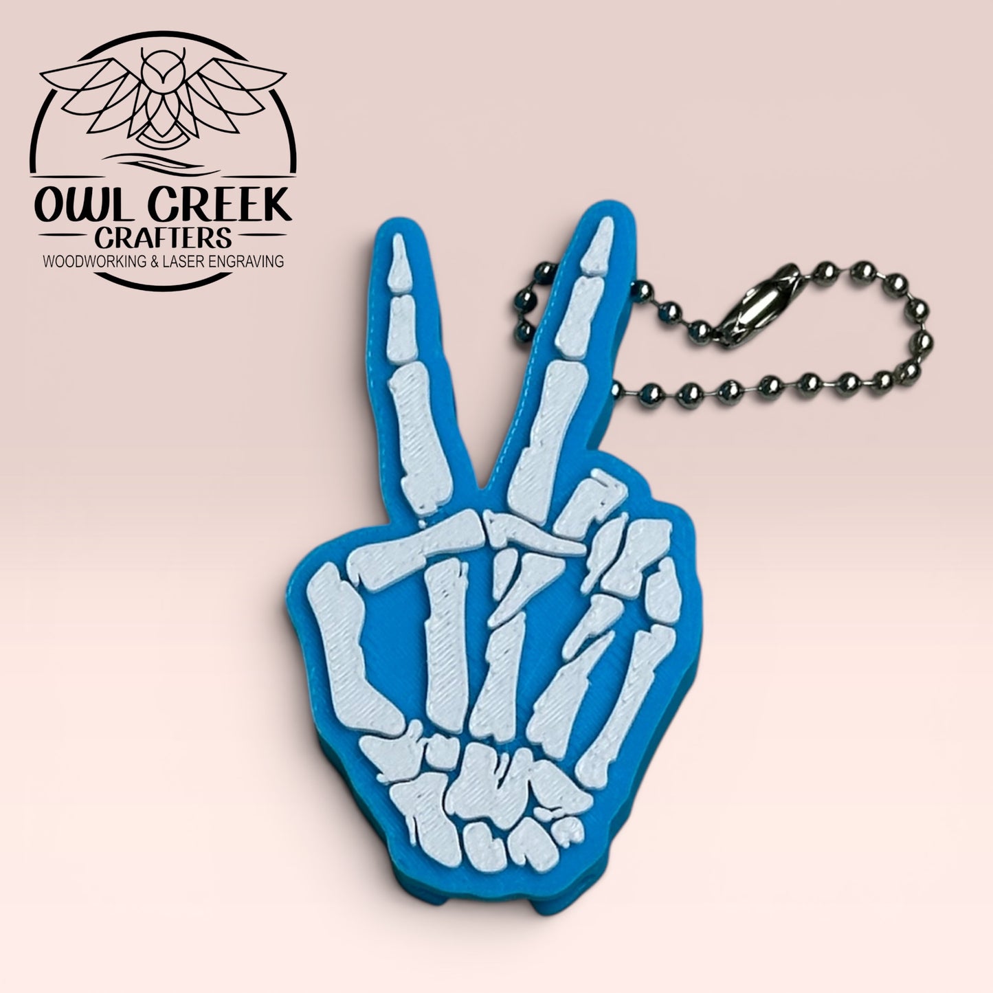 Blue and white skeleton hand keychain making a peace sign on a beige background with Owl Creek Crafters Logo