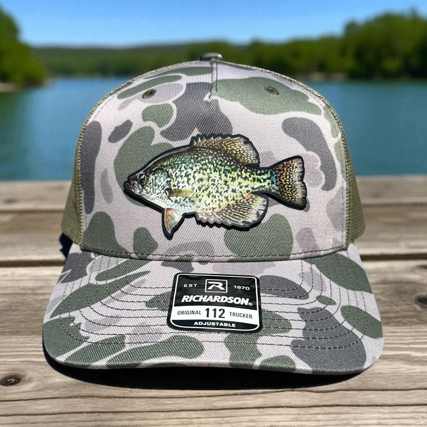 Blue and green camouflage trucker cap with uv printed fish design patch on a wooden surface with a lake background