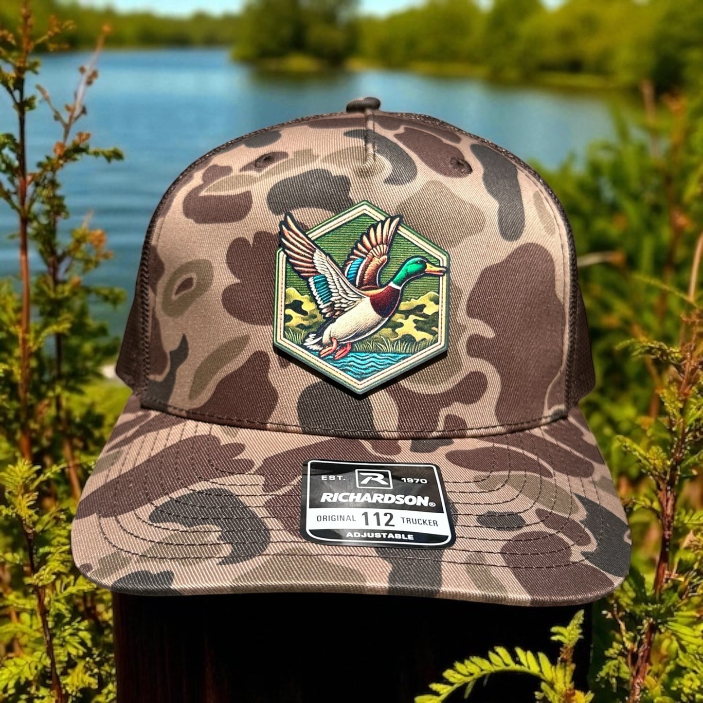Brown camouflage trucker cap with a hexagon uv printed mallard duck patch and visible brand logo label against a natural lake background