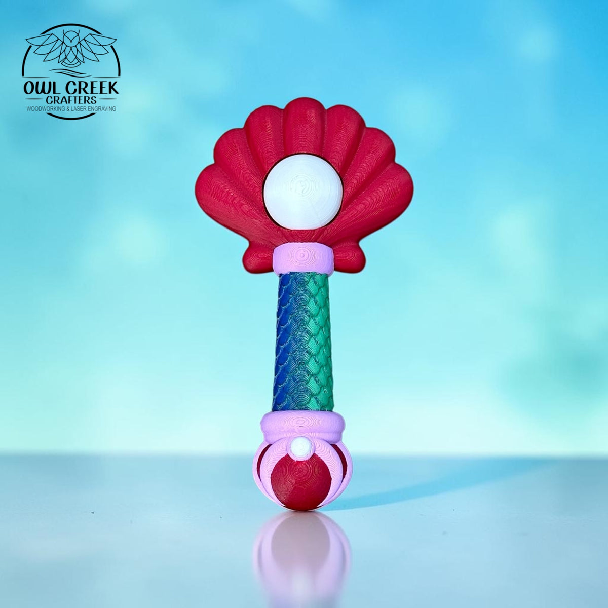A 3d printed wand with a red sea shell top, a green and blue scaled wand handle, with purple accents. There is a "pearl" in the middle of the sea shell that clicks. On a blue and white sea background with the 'Owl Creek Crafters' Logo in the top left corner.