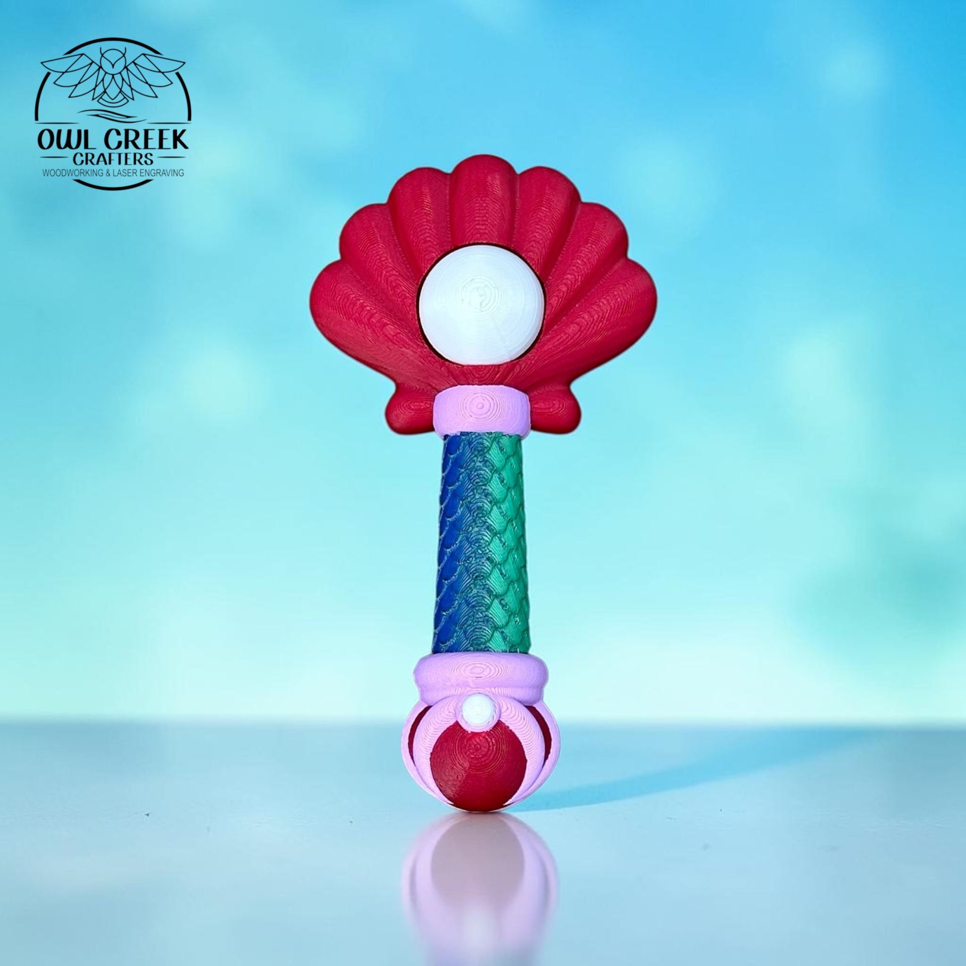A 3d printed wand with a red sea shell top, a green and blue scaled wand handle, with purple accents. There is a "pearl" in the middle of the sea shell that clicks. On a blue and white sea background with the 'Owl Creek Crafters' Logo in the top left corner.