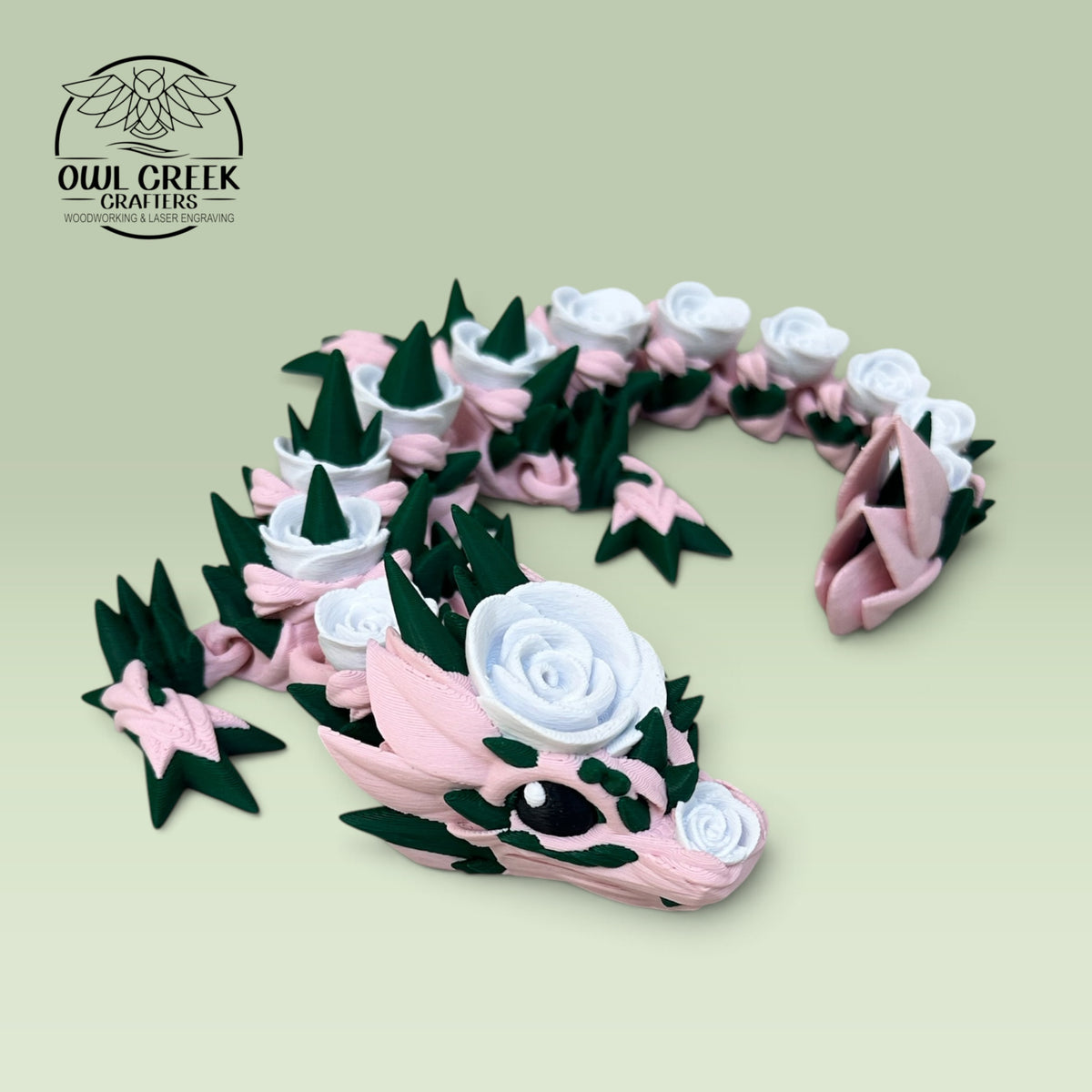 A 3D-printed articulated dragon figurine in soft pink and dark green, decorated with white rose details, displayed on a light green background. The Owl Creek Crafters logo is visible in the top-left corner.